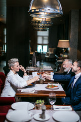 Satisfied cheerful business partners in formal clothes, sit at setted table, raise wine glasses up with joyful faces, passionately celebrate birthday of charming colleague, background of big window