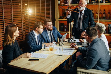 Bearded man in expensive suit and nice tie shows mock helicopter to team of young partners, pretty female in black suit watching executive director attentevely.