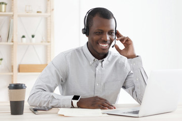 Smiling african customer support service operator with hands-free headset working in office, using laptop