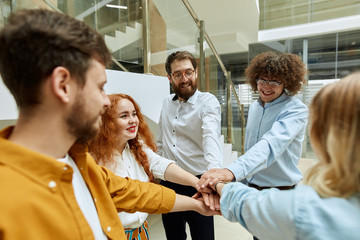 Joyful photo of young cheerful team of talented designers standing in modern office, happily keeping hands together, showing unity and friendship, indoor shot