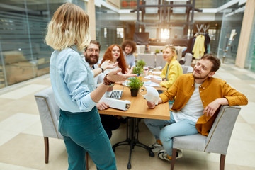 Young blonde woman with wavy hair stands self confidently back to camera, spreads hands sideways explaining bright business idea, playful colleagues listen to her attentively.