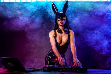 Young sexy woman dj playing music in mask. Headphones and dj mixer on table. Smoke on background