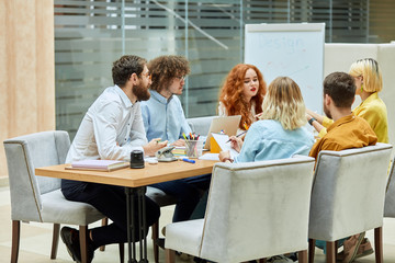 Joyful young colleagues, casually dressed, discuss new design project, sitting at wooden table in fashionable gray armchairs, busy at work, left side shot