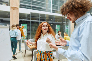 Pretty foxy haired woman dressed in white blouse, spreads hands sideways, suspiciously looking at kind partner with Afro hairstyle in modern designers agency