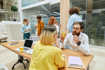 Obraz premium Handsome polite designer, wears round spectacles, put elbows on working wooden table, passionately speaks with female partner sitting opposite him at table, background of crowded office