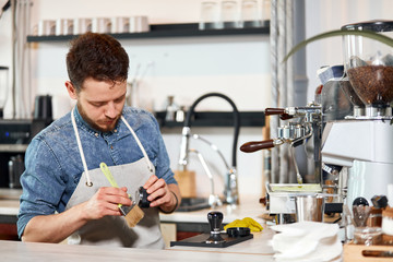 Skillful profesional male barista, stands near coffee maker, cleans working place, looks down, examins metal tamper , working hard in modern restaurant