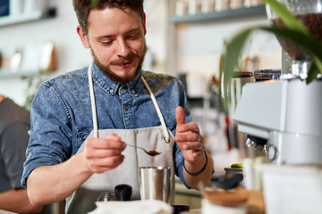 Portrait of young cheerful man working at counter, holding silvel spoon with ground coffee ready to put it into metal coffee pot in brightly lighted room
