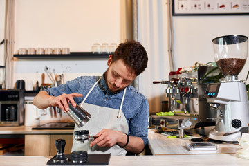 Mindful calm barista with thick beard, wears apron, bows head, looks away, puts coffee into tamper, works in modern coffee house