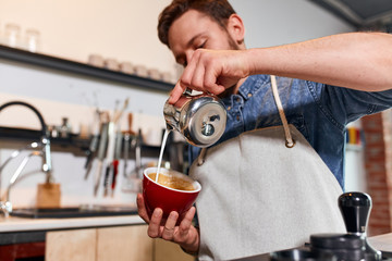 Handsome professional barista preparing cup of fresh hot latte for friendly customer in coffee shop, working time concept