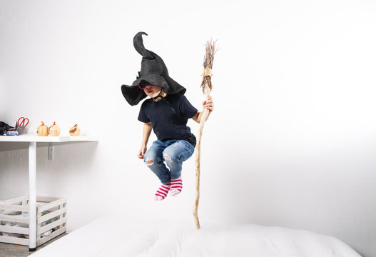 Boy in a witch hat jumps on a mattress with a broom.