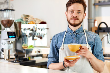 Fototapeta na wymiar Professional young barista stands with yellow coffee cut, looks at camera with calm face, demonstrates thankful, ready to give fresh prepared latte to customer, coffee house concept