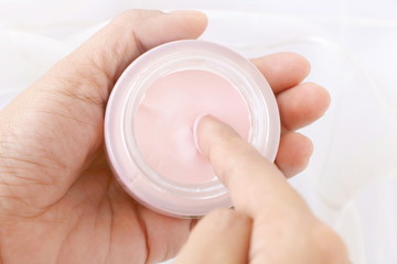 Top view of close up woman finger applying pink cream in jar on white fabric background