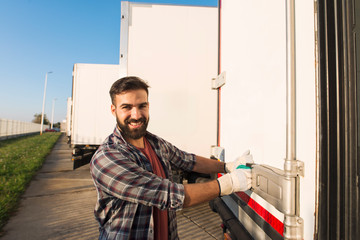 Smiling truck driver in working gloves opening or closing truck trailer back doors checking goods...