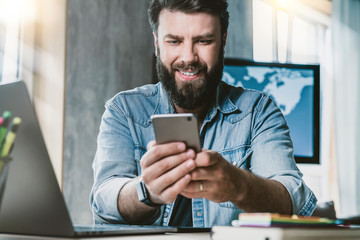 Man sitting at desk and holding mobile phone in hand. Hipster guy watching funny videos on mobile device and smiling.