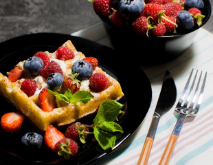 Belgian waffles with raspberries, strawberries and blueberries on a black plate. Selective focus.