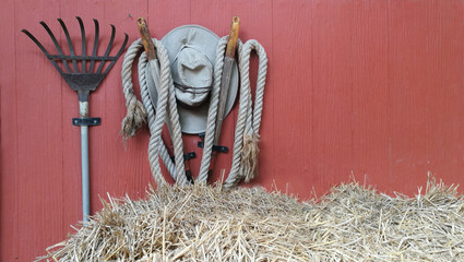farming instrument like rake and hat hang on wall with straw on floor.