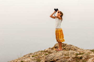 Child girl traveler stands on a rock above the water and looks through binoculars up into the sky. T-shirt and colored yellow skirt.