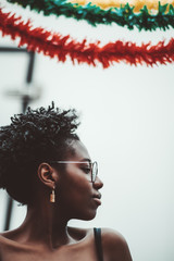 A vertical portrait of a young charming African female in spectacles and with earrings, she is looking aside, three garlands above of red, green and yellow colors; a dazzling black girl outdoors