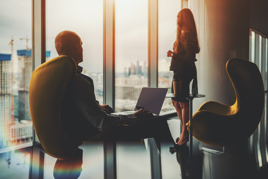 Two Silhouettes Of Business Partners On The Top Floor Of A Luxurious Office Skyscraper In Front Of The Window With A Construction Site Outside: Man Entrepreneur With A Laptop And His Female Colleague