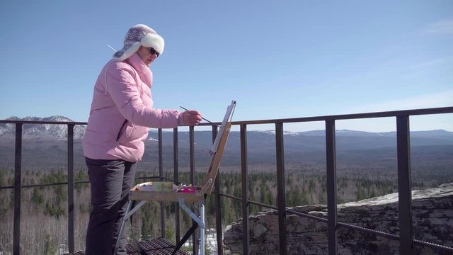 Grandmother at the peak of a winter mountain, paints a picture in oil. Woman and malbert outdoors in the new year.