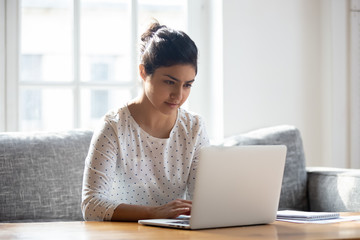 Focused Indian woman using laptop at home, looking at screen