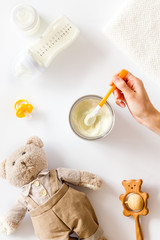 Make mixture for feed baby. Spoon with food in hand near toys and accessories on white background top view pattern