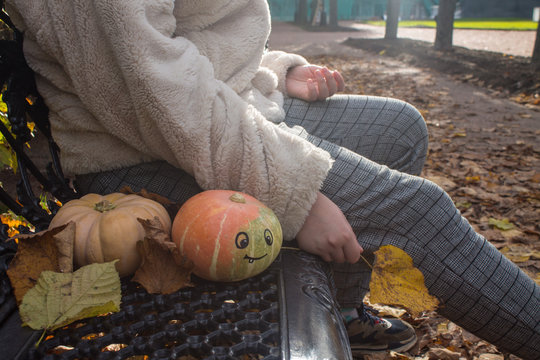 A pumpkin with a funny painted face on a bench and a teenage girl with an autumn leaf in her hands