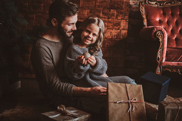 Cheerful little girl is holding christmas decoration ball while sitting with her father on the floor, surrounded with gifts.