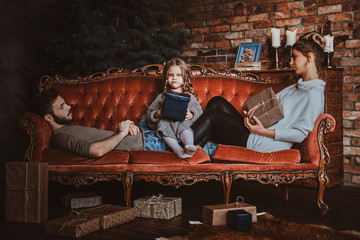 Young little family are chilling on red vintage sofa with presents in hands.