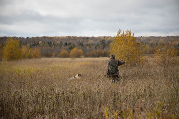 The hunter is walking across the field, with weapons. Next to the dog is a springer spaniel. Autumn