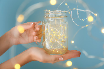 Female hands with glowing garland in jar on color background