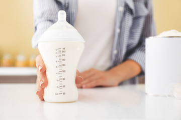 Mother with bottle of baby milk formula on table in kitchen, closeup