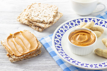 Creamy peanut butter. Paste in bowl, sandwich and peanuts on white wooden table.