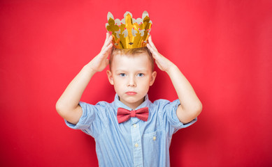 Happy education and childhood concepts with an adorable 6-year old boy holding a golden king crown on his head as a wise spoiled child, red background