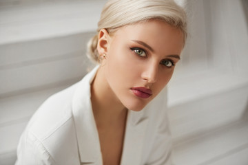 A fashionable young woman with perfect blond hair and perfect trendy makeup in an elegant white suit posing in studio. Beautiful blonde model girl with red full lips and deep seductive eyes.