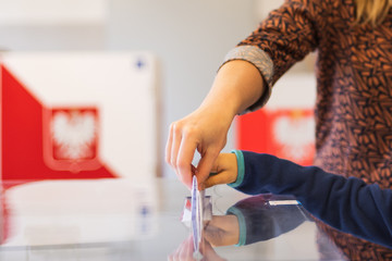 Woman's and kid's hands puting a card with a vote.to the ballot box during parliamentary elections. In the backround polish arms and flag - obrazy, fototapety, plakaty