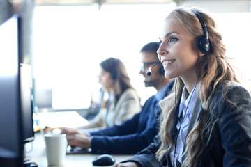 Female customer support operator with headset and smiling, with collegues at background.