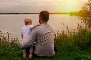Fototapeta premium Daughter with father on the shore at sunset. Autumn, view from the back. Fatherhood and care.