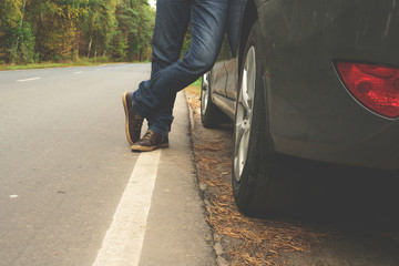 A young man is standing near a gray car on an empty road, on an empty highway. Legs of a man near a car on the highway. Lifestyle.