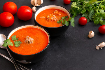 black plates of red tomato soup with meat meatballs, greens on a black background, spoons, garlic, fresh cherry tomatoes,