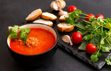 black plate of red tomato soup with meat meatballs, greens on a black background, garlic, fresh cherry tomatoes,
