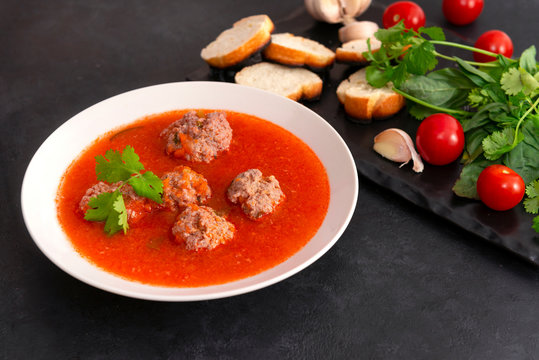 1 white plate of red tomato soup with meatballs, greens on a black background, garlic, cherry tomatoes, cilantro, Basil