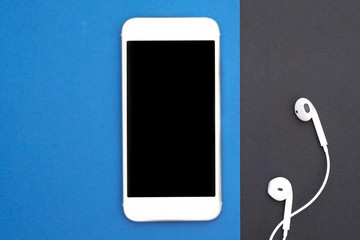 Music, gadgets, music lover. White smartphone on the black and blue background with headphones. View from above. Flat lay, top view.