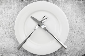 Table setting. Empty plate, knife and fork on a light concrete background. The fork and knife lie crosswise on a plate, meal is finished, the dish was not pleasant. Top view, flat lay, copy space.