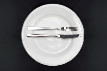 Table setting. Empty plate, knife and fork on a black background. The fork and knife lie on a plate in parallel, the meal is finished, I liked the dish. Top view and flat lay with copy space