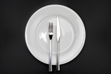 Table setting. Empty plate, knife and fork on a black background. The fork and knife lie on a plate in parallel, the meal is finished, I liked the dish. Top view and flat lay with copy space
