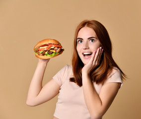 Young red-haired girl holds a burger sandwich, is surprised, on a beige background