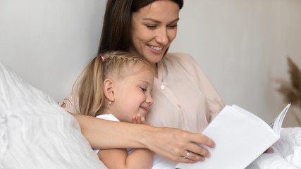 Obraz na płótnie Canvas Smiling mother reading book to cute little daughter in bed
