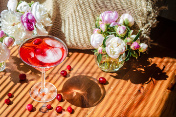 Solemn rustic still life with glass of red drink with autumn berries and ice surrounded by white flowers on textile background. Wooden table in the striped shadow of the blinds on bright sunny day