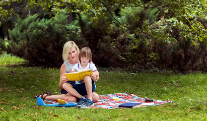 Young mother reading a book with her special son on a rug amidst a park lawn